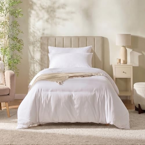 A neatly made bed with white bedding and a light gray tufted headboard in a softly styled bedroom. A beige nightstand with a lamp, a chair, and a plant are visible around the bed.