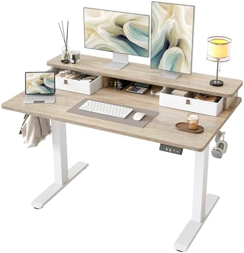 A modern standing desk with a light wood grain top and white T-style legs, shown with a computer monitor, laptop, keyboard, mouse, lamp, and storage drawers on a clean white background.