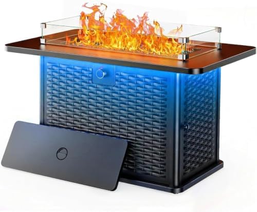 A modern outdoor fire pit table with a black wicker-patterned body, dark brown tabletop, clear glass wind guard, and active orange flames. A black control panel is shown leaning against the front left side on a white background.