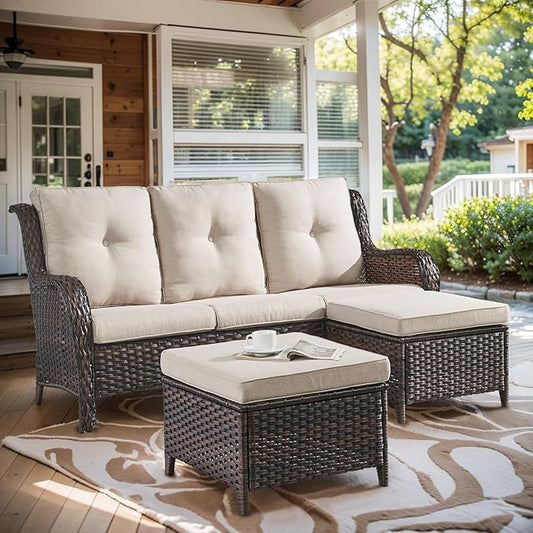 A three-seat wicker patio sofa with tufted cream cushions, a matching chaise ottoman, and a small square coffee table, arranged on a patterned rug on a covered porch with outdoor greenery visible in the background.