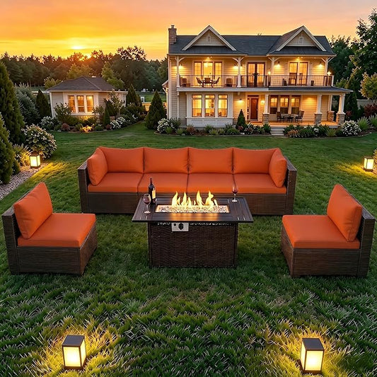 Outdoor patio furniture set on a grassy lawn in front of a two-story house at sunset, featuring a dark brown wicker-style sectional sofa, two matching armchairs, and a square fire pit table with visible flames.